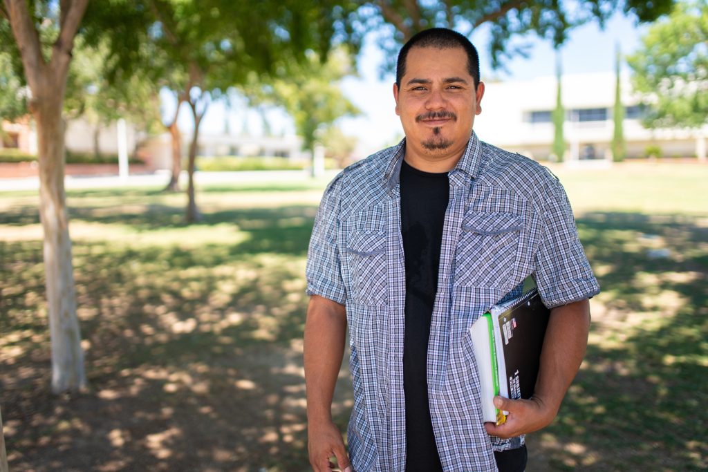 Jesus Serrano, a smiling Latino man with a blue plaid shirt and school books under his arm, who was able to earn his GED with help from The Job Spot.