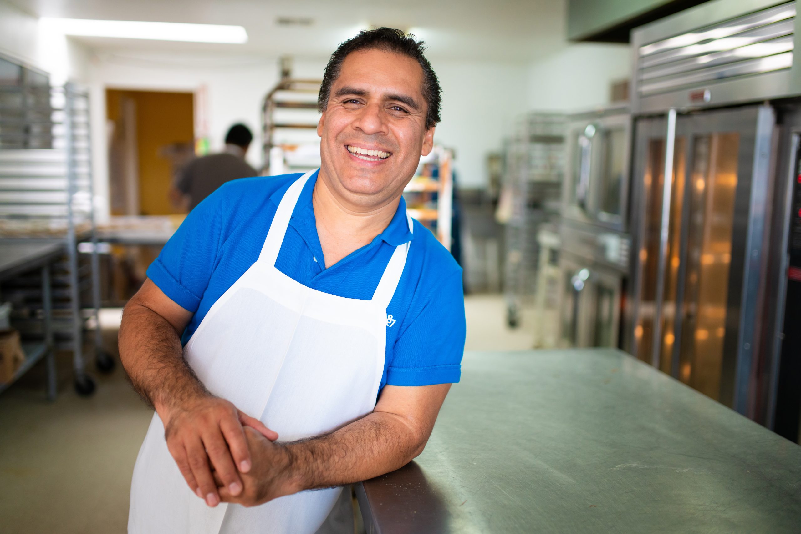 Juan Rodriguez, a cheerful middle-aged Latino man in a baker's apron, smiles for the camera in his bakery's kitchen.