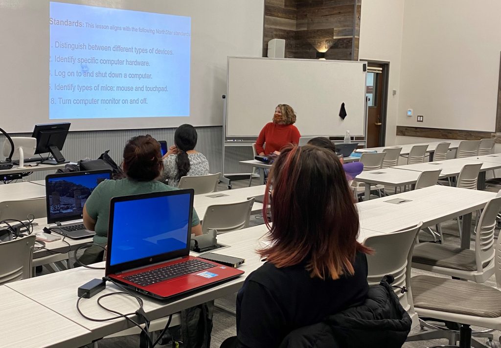 Several adult students sit in a classroom at long desks with their laptops while watching their teacher's powerpoint presentation to help them become job ready.