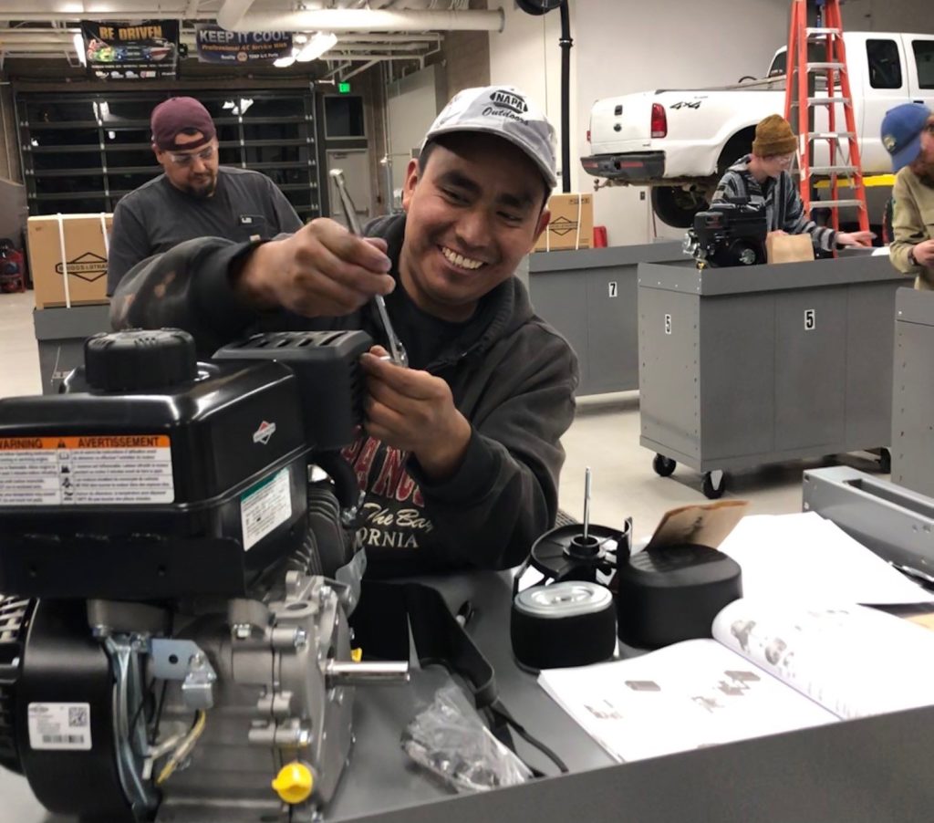 A smiling man works on an engine in a mechanic's shop