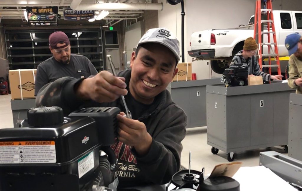 A smiling man works on an engine in a mechanic's shop