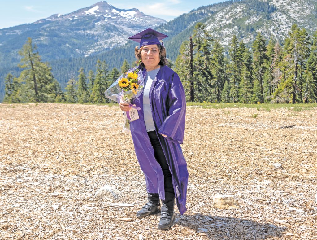 Meli Aguirre, a middle aged woman, stands in front of a line of evergreen trees and mountains in a purple graduation cap and gown, holding a bouquet of sunflowers.