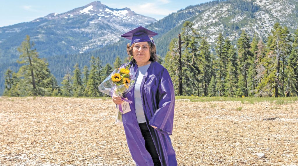 Meli Aguirre, a middle aged woman, stands in front of a line of evergreen trees and mountains in a purple graduation cap and gown, holding a bouquet of sunflowers.