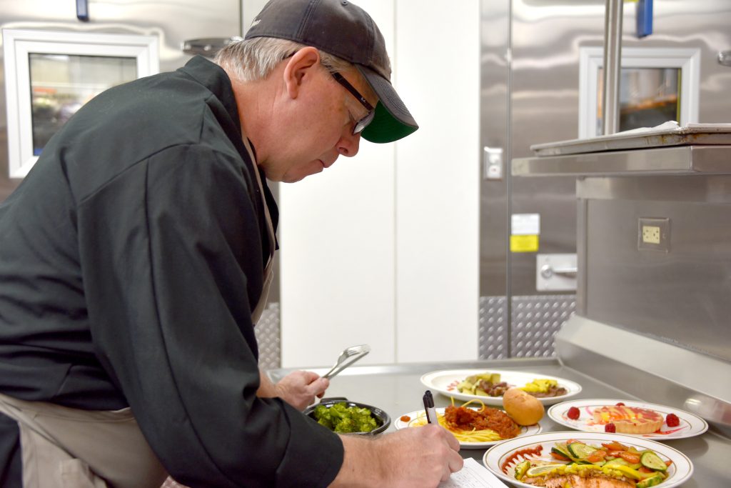 David Smith, and older man in a black uniform and ball cap, leans over several plates of food in a professional kitchen to check the work of culinary students.