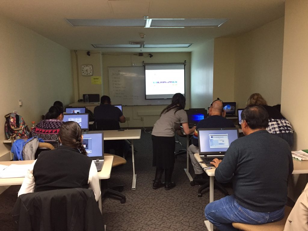 Two rows of adult students sit in a small classroom with laptops, looking at their teacher's lesson projected on the wall.