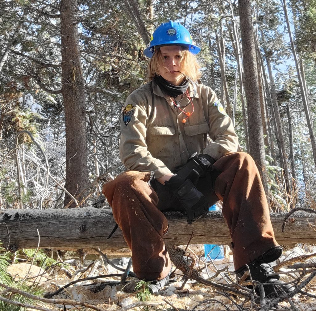 Riley Hinkson, a blonde woman in forestry work gear, sits on a log in a forest after building her new career thanks to ADVANCE's whole-person approach.