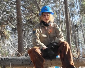 Riley Hinkson, a blonde woman in forestry work gear, sits on a log in a forest.