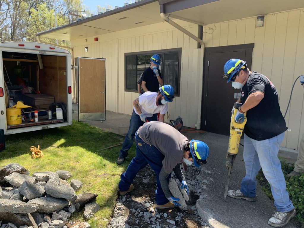 A group of adult students doing construction work to break up a concrete pathway in front of a building.