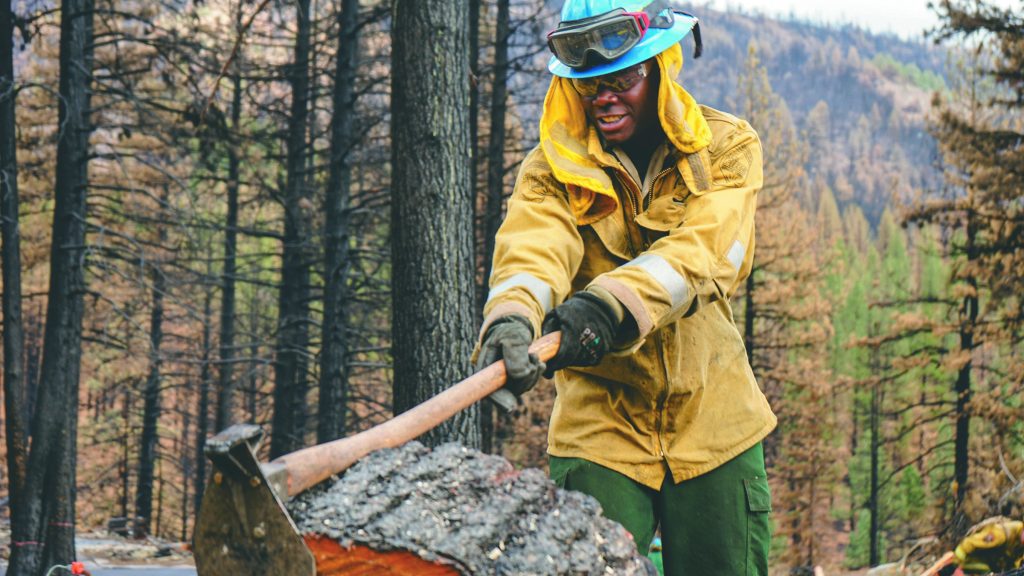 A black man in a yellow work jacket and blue hard hat chops at a large log with an ax.