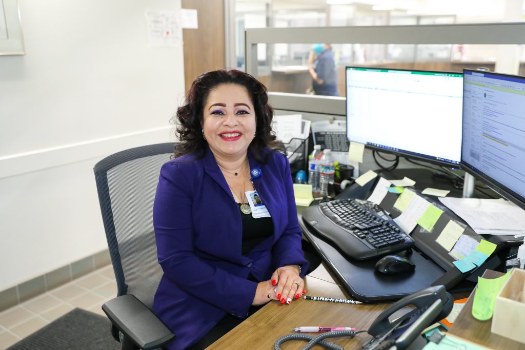 Yesenia Quintanilla, a smiling middle aged woman in a sapphire blue blazer, sits at her office desk.