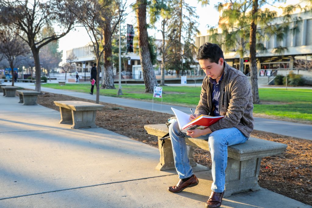 Juan Carlos Romera, a young man in a grey jacket and jeans, sits on a bench at his school campus writing in a notebook.