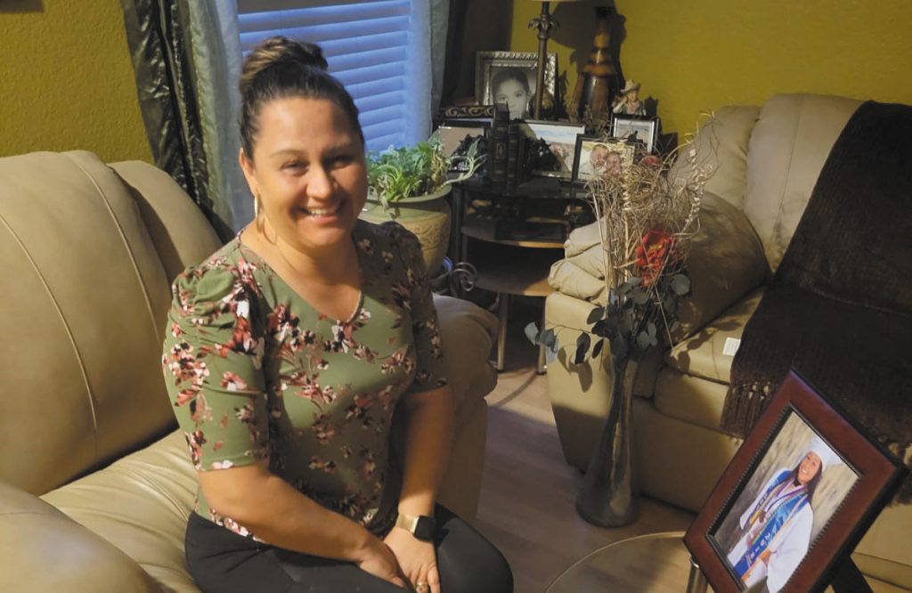 Maria Villatoro site on an armchair in her home in front of a photo of herself in her graduation cap and gown and smiles for the camera.