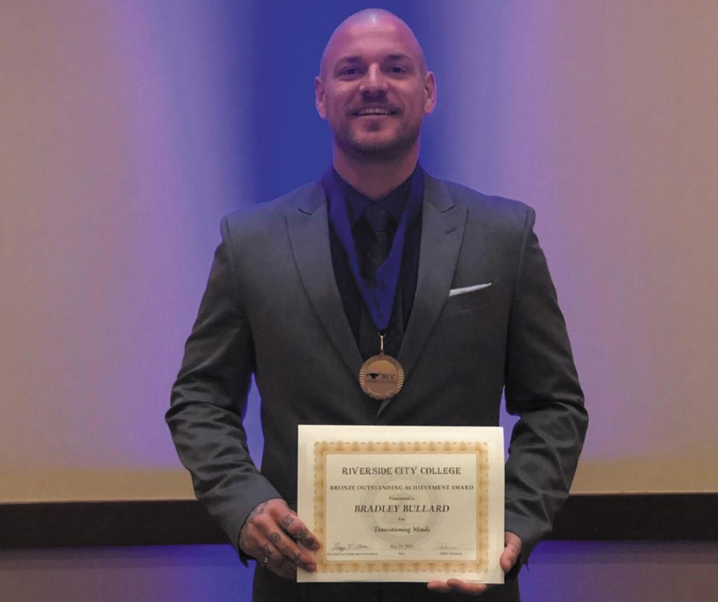 Bradley Bullard, a man in a nice charcoal grey suit, stands holding his diploma and wearing a medal.