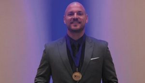 Bradley Bullard, a man in a nice charcoal grey suit, stands holding his diploma and wearing a medal.