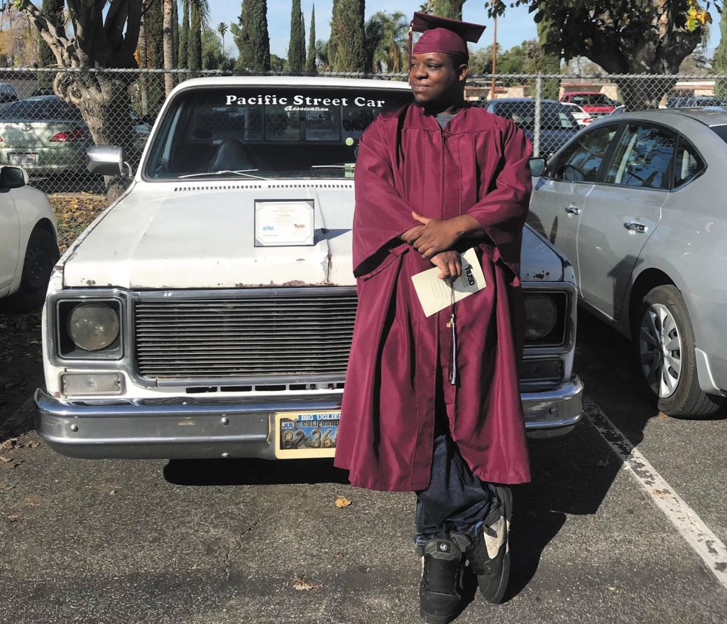 Jason Merriweather, a black man wearing a dark maroon graduation cap and gown, leans against a white classic car. 