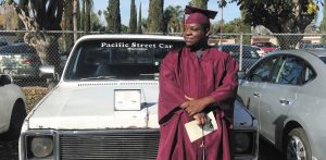 Jason Merriweather, a black man wearing a dark maroon graduation cap and gown, leans against a white classic car.