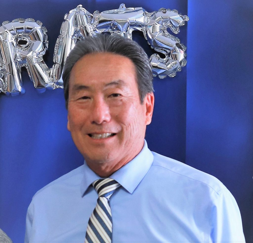 Rich Ishimaru, an older Asian man in a blue collared shirt and striped tie, smiles for the camera.