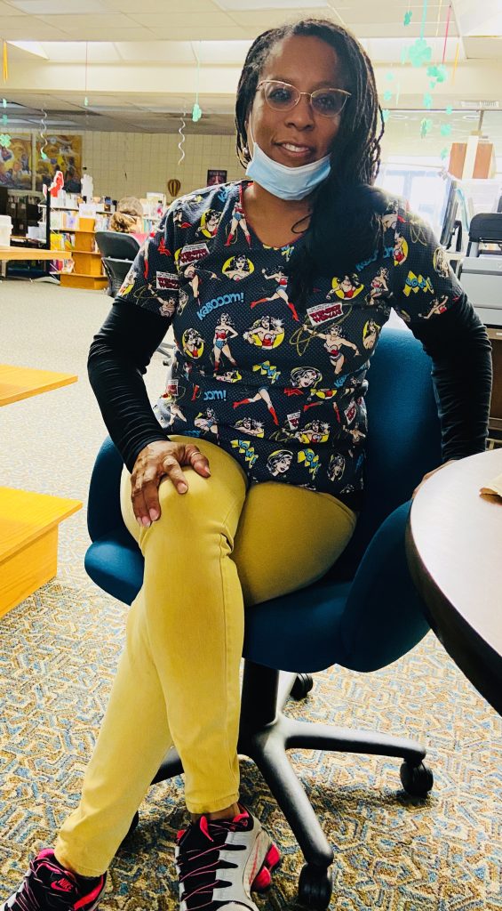 Joyce Glaspie, a black woman wearing cat eye glasses, superhero scrubs, and a lowered disposable face mask, sits in an office chair and smiles for the camera.