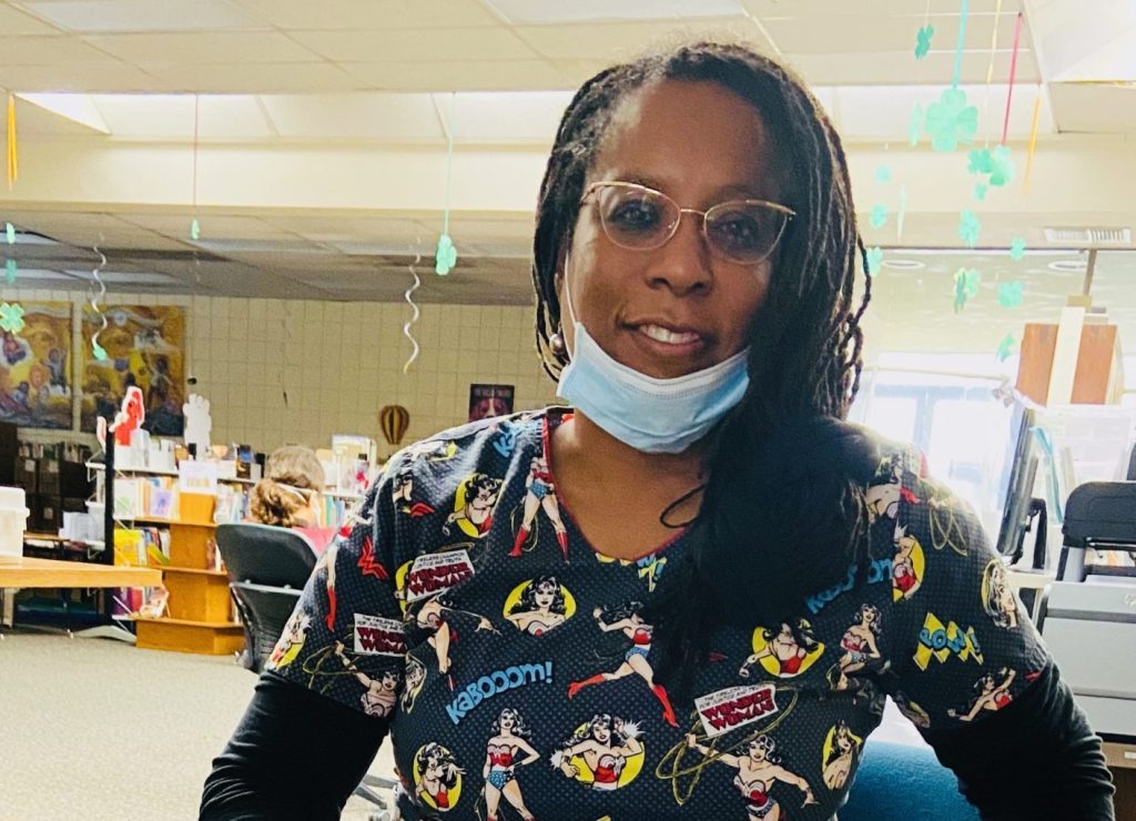 Joyce Glaspie, a black woman wearing cat eye glasses, superhero scrubs, and a lowered disposable face mask, sits in an office chair and smiles for the camera.