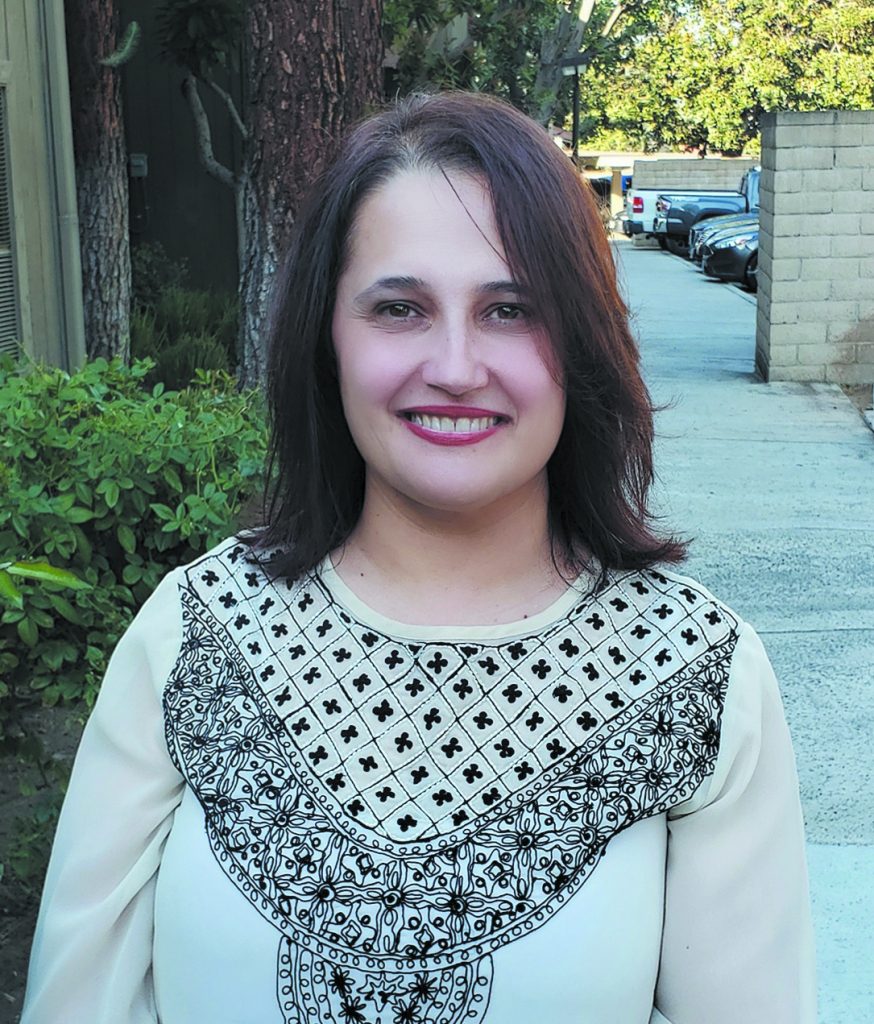 Luma Dawood, a woman in a white and black patterned blouse and shoulder length reddish-brown hair, smiles for the camera in a yard.