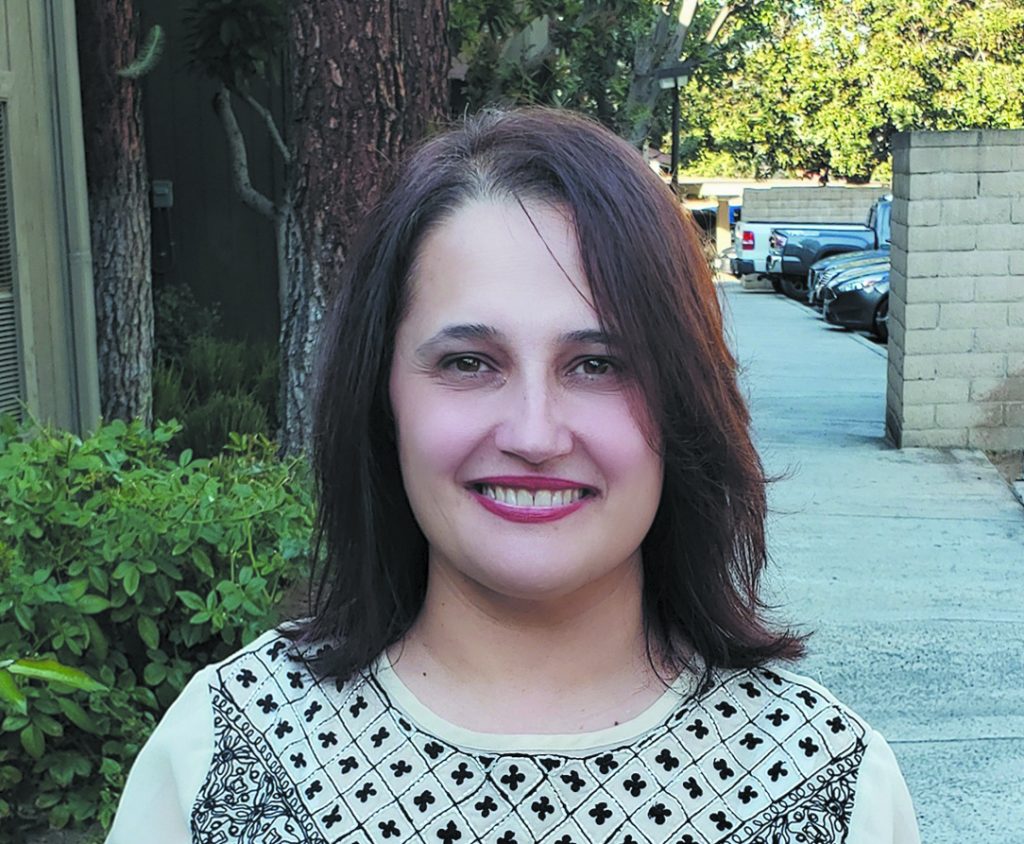 Luma Dawood, a woman in a white and black patterned blouse and shoulder length reddish-brown hair, smiles for the camera in a yard.