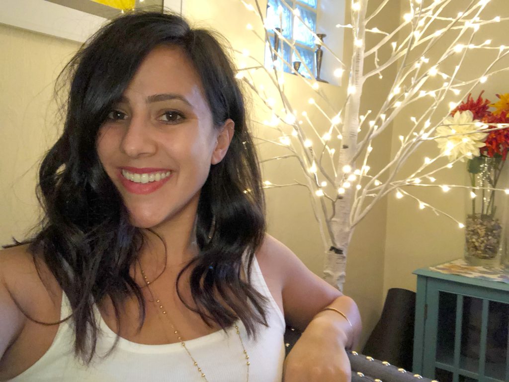 Shireen Franco, a young dark haired woman in a white tank top, smiles for the camera in her home.