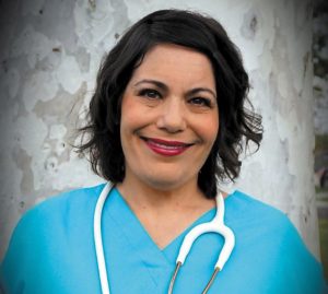 Alicia Torres-Wynne, a woman with short dark hair, blue scrubs, and a white stethoscope around her neck, smiles for the camera.