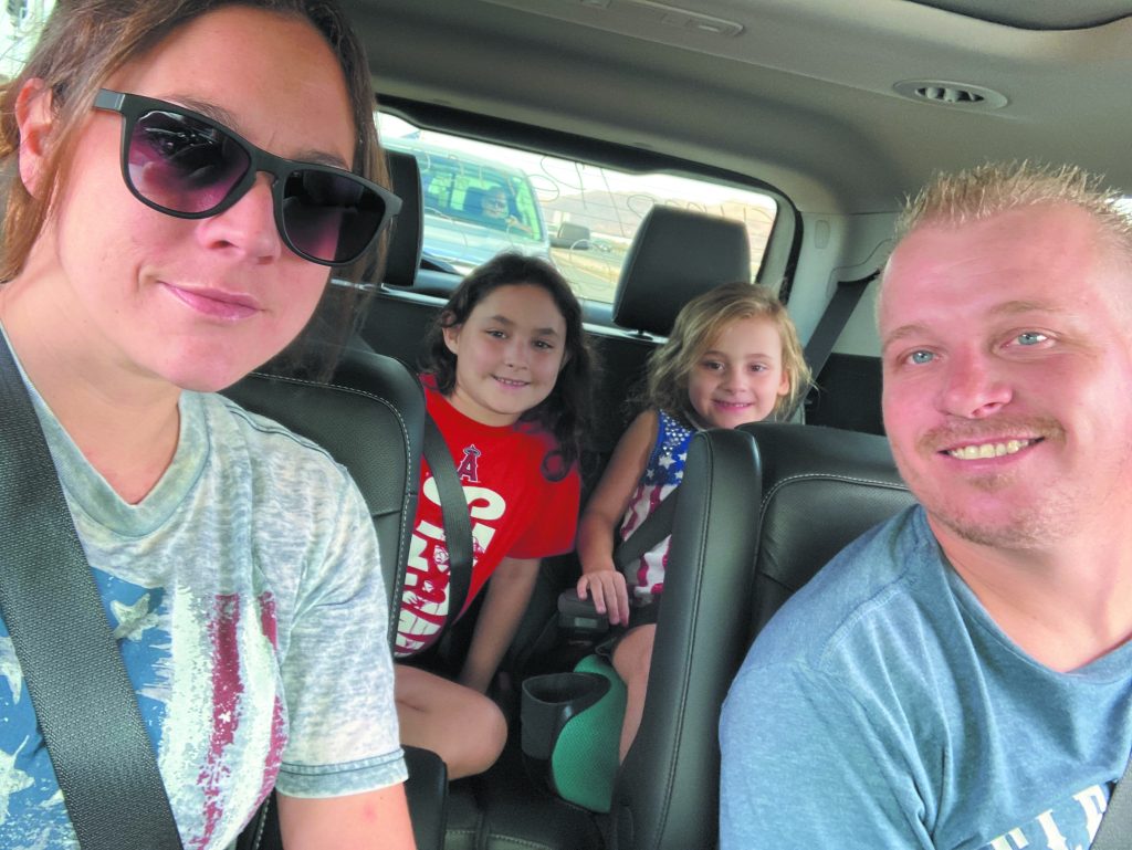 Krysti Lopez sits in the passenger's side of a car in a grey t-shirt and sunglasses, taking a selfie. Her husband, a blond man in a blue shirt, learn into camera view, and her two young children, a boy and a girl, smile from the back seat.
