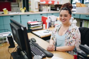 A woman sits at a desk, smiling and holding her hands in front of her chest. She is in an office, and a desktop computer is in front of her.