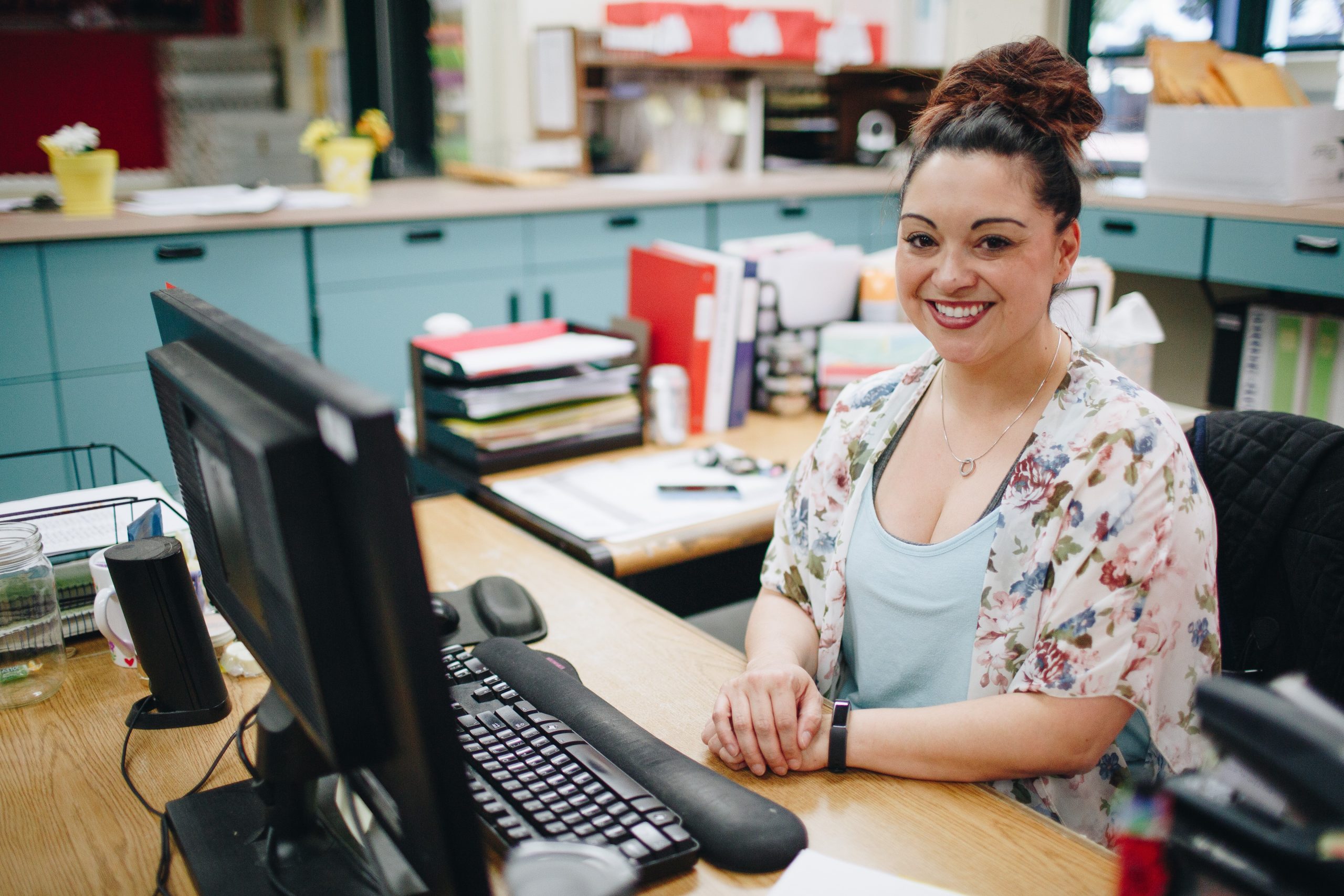 A woman sits at a desk, smiling and holding her hands in front of her chest. She is in an office, and a desktop computer is in front of her. She is at her dream job.