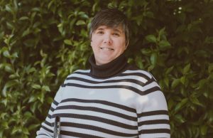 Janna Evans, a woman with short hair and a white sweater with black stripes, stands in front of foliage and smiles at the camera.