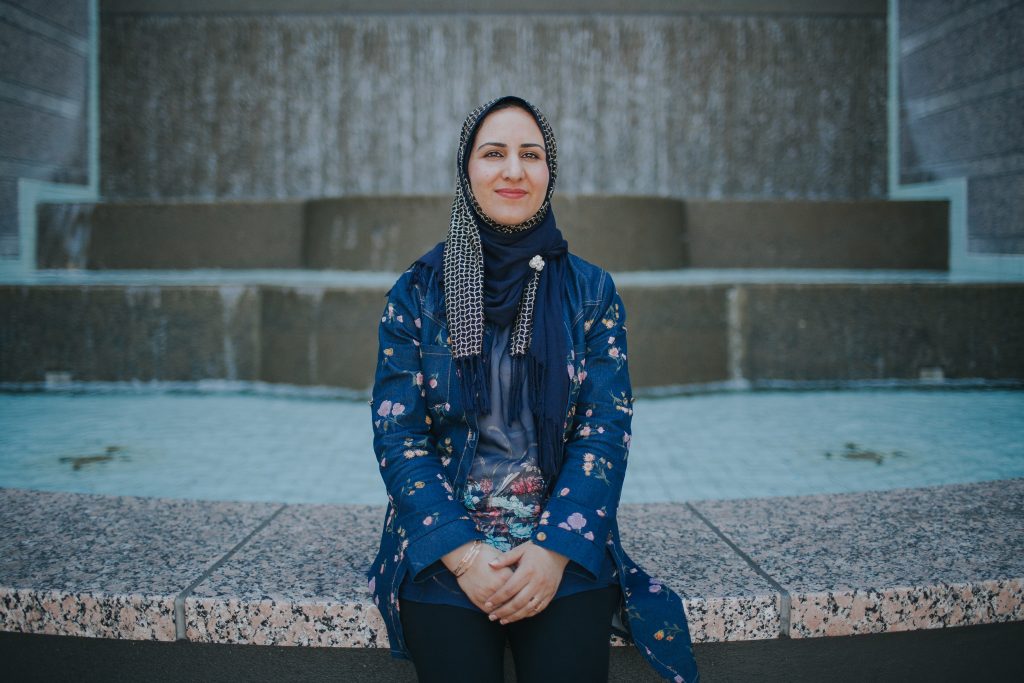 Hamida Hakimi Roshangar, a woman in a hijab and blue patterned blouse, sits on the steps of her adult school campus.