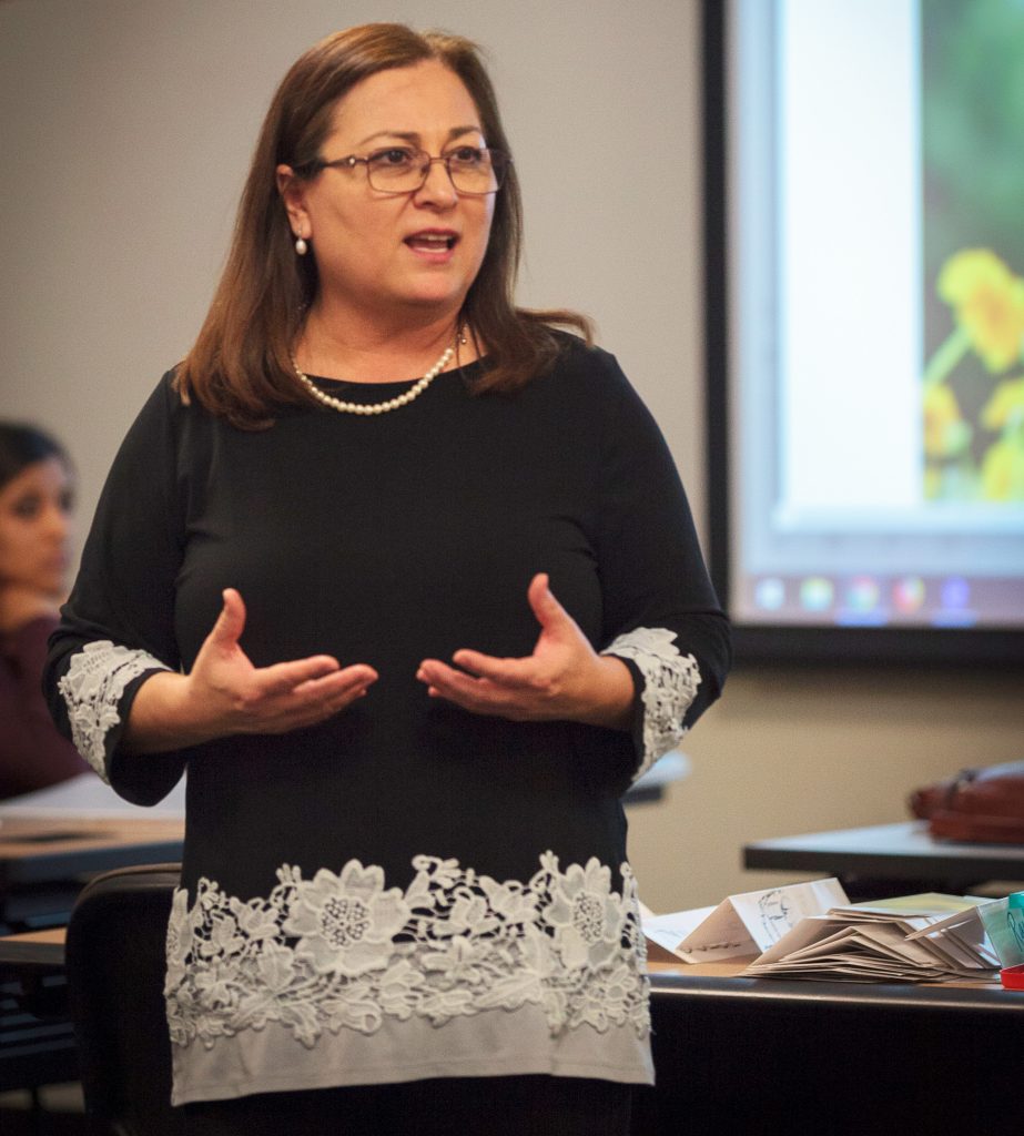 Irma Gonzalez, a woman in a black blouse with white lace detailing, lectures in a classroom.