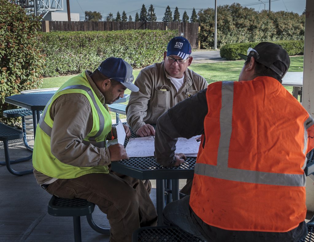 Three men in construction gear sit around a table outside looking at work papers