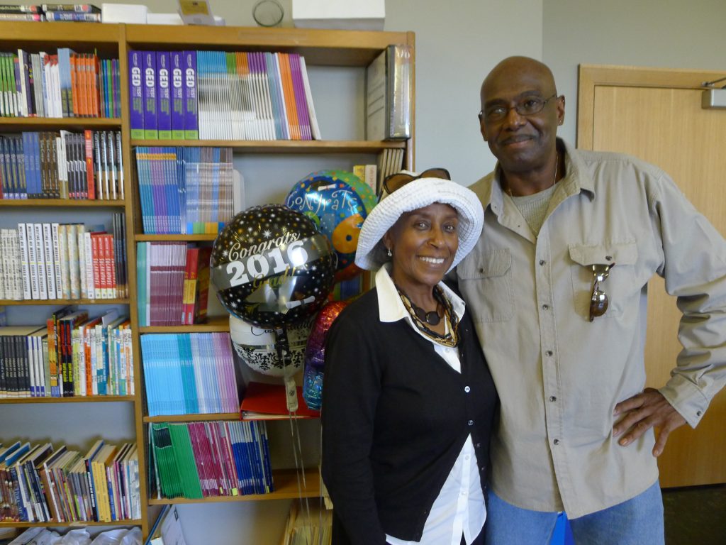 A man who participated in the Second Start Adult Literacy Program and woman stand in front of a book shelf. There a balloons next to them, one saying "Congrats Grad 2016."