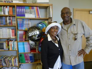 A man and woman stand in front of a book shelf. There a balloons next to them, one saying "Congrats Grad 2016."