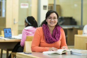 Una mujer joven sonríe mientras está sentada en un escritorio en una biblioteca. Otra mujer está sentada detrás de ella con una computadora portátil abierta.