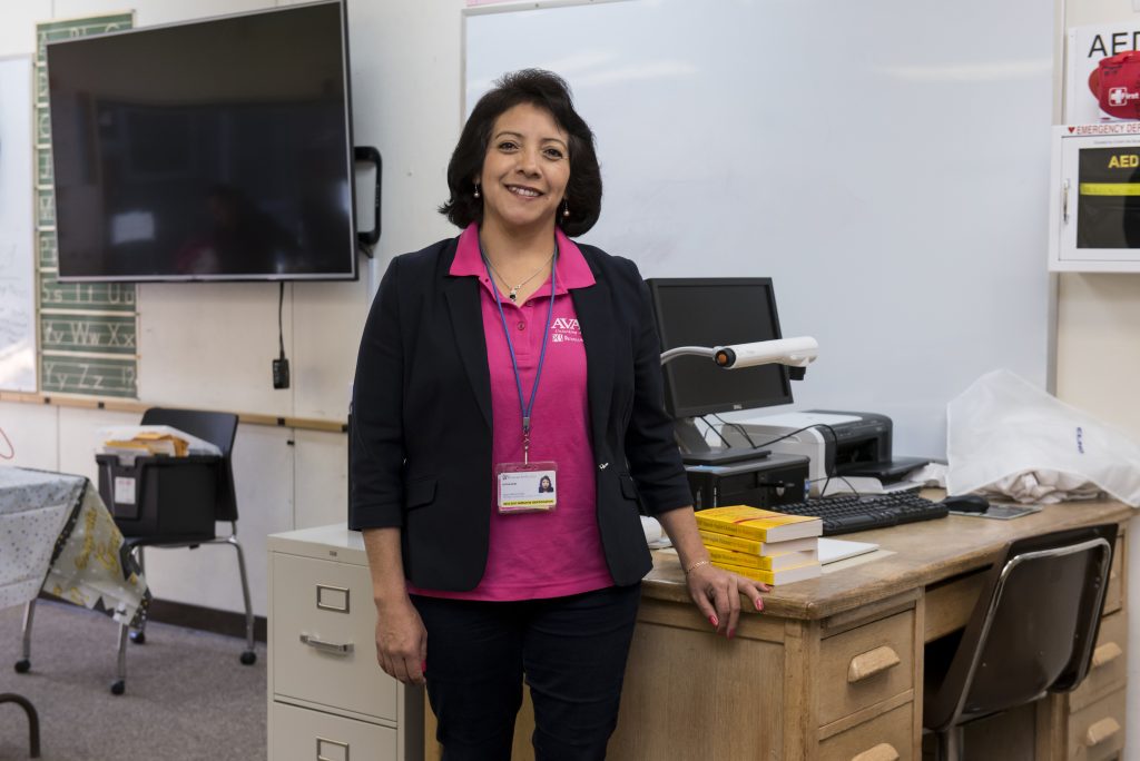 A woman stands in front of desk in a classroom. She is smiling. There is a mounted TV behind her.