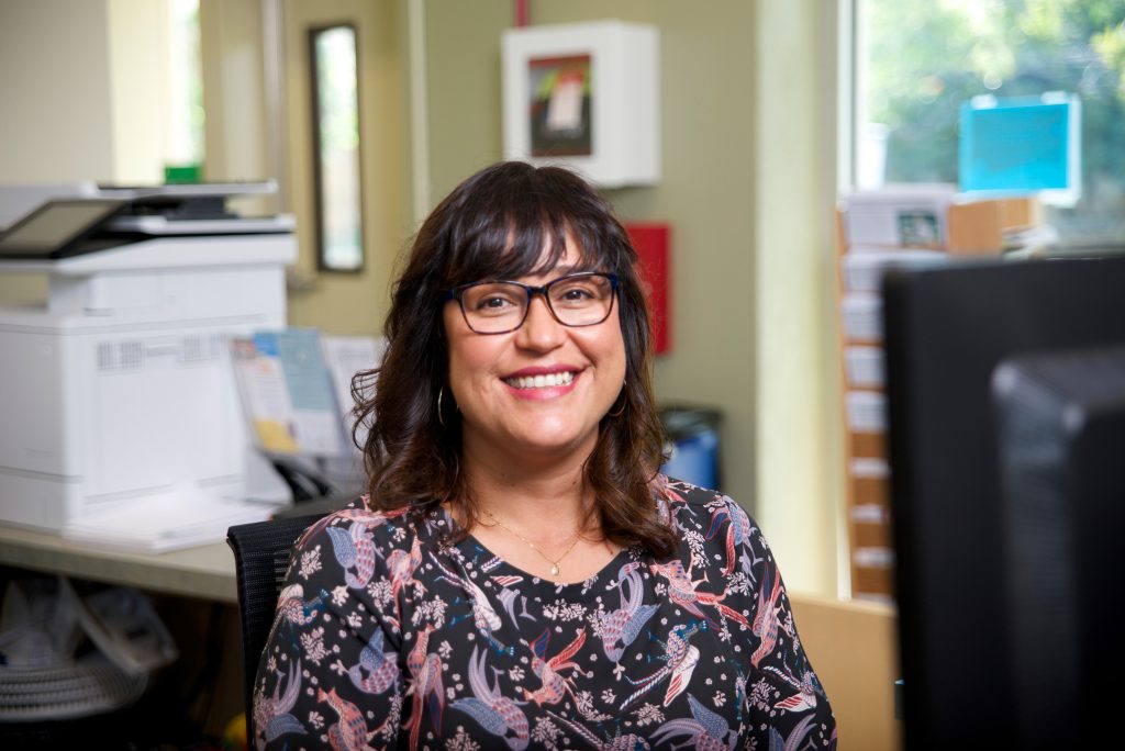 Olga Saucedo sits at her desk and smiles for the camera.