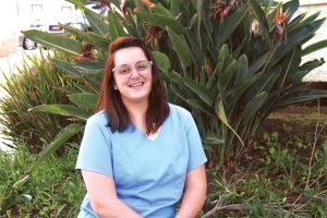 Autumn Diaz, a young woman with red toned hair, glasses, and blue scrubs, sits outside in front of large plants.