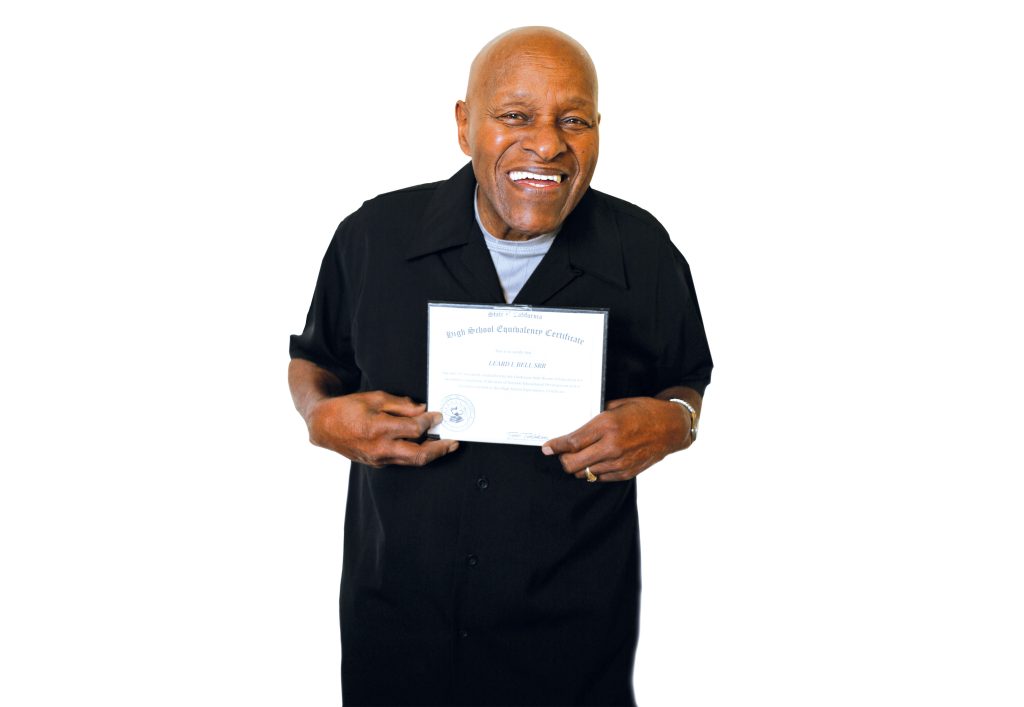 Leard Bell, and elderly black man in a black button up shirt, holds his GED certificate/