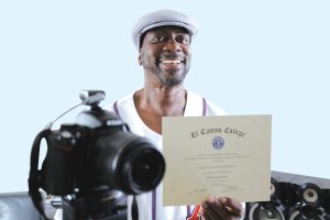 Vincent Meredith, a black man in a flat cap, stands with his photography gear and adult school certificate.