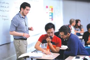 An adult school teacher in a button up shirt and slacks stands over two students, watching them work in class.