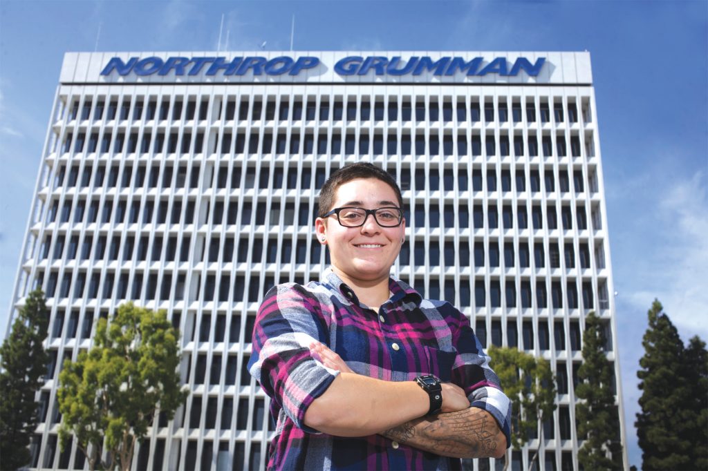 Amanda Gonzalez in a plaid shirt and glasses stands in front of the Northrop Grumman building where she works.