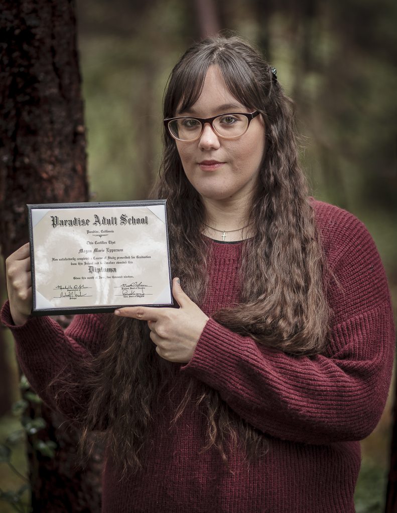 Meagan Epperson, a woman with cat eye glasses and a dark red sweater, holds her high school diploma.