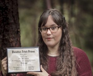 Meagan Epperson, a woman with cat eye glasses and a dark red sweater, holds her high school diploma.