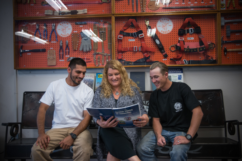 Staci Teegarden and two students sit in front of a wall of tool looking at a book about iron working.