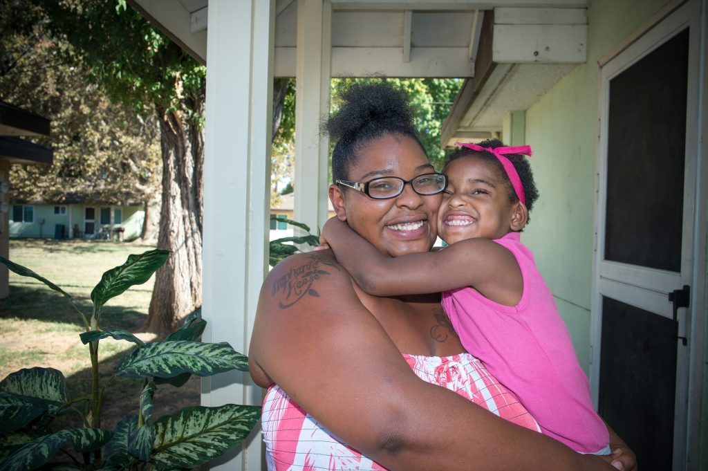 Katerria Guichard, a black woman in glasses, and her young daughter, both wearing pink, stand on the porch of their home.