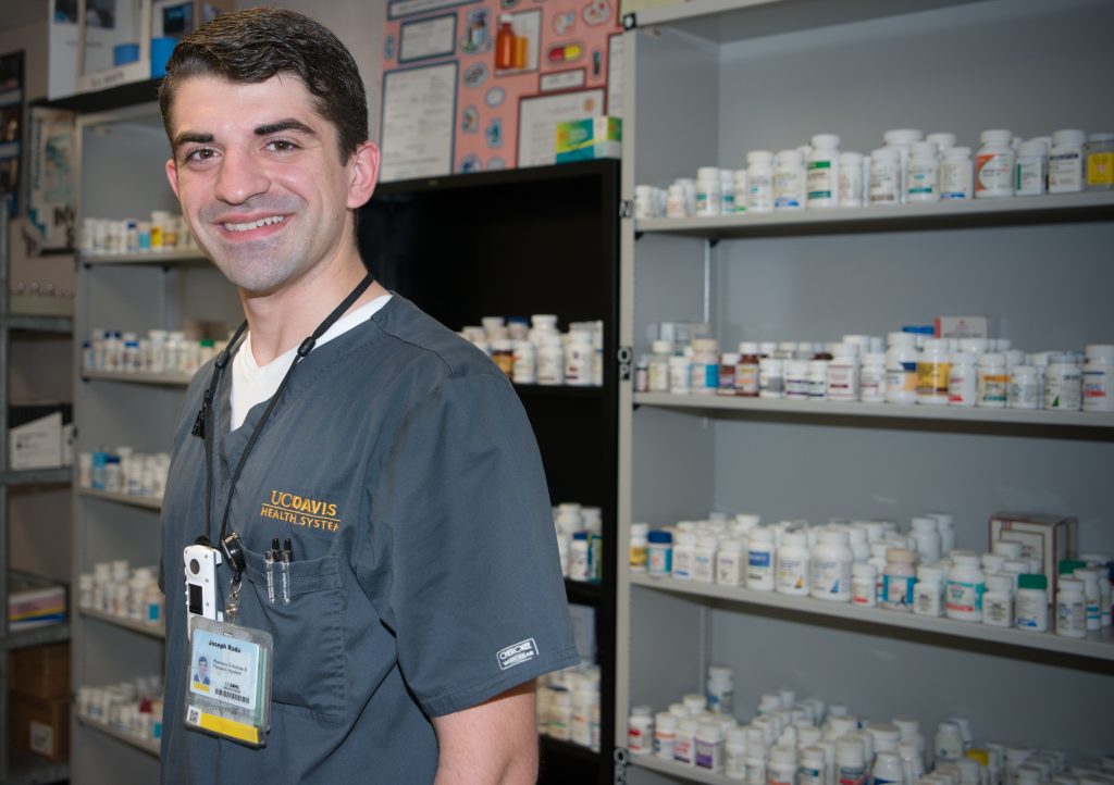 Joseph Radu, a white man in dark grey UC Davis scrubs, stands in front of shelves of medication bottles at his place of work.