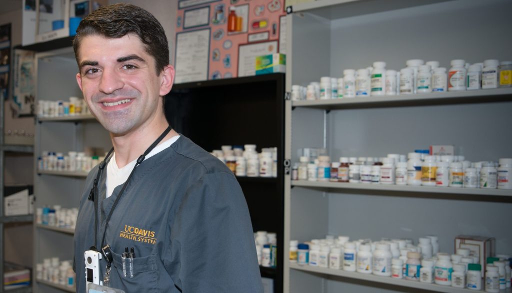 Joseph Radu, a white man in dark grey UC Davis scrubs, stands in front of shelves of medication bottles at his place of work.